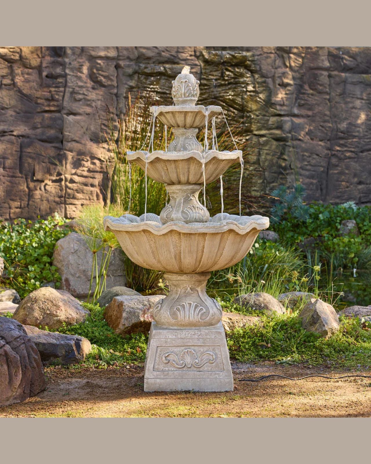 Fleur three-tiered stone fountain in a garden setting with rocks and plants.