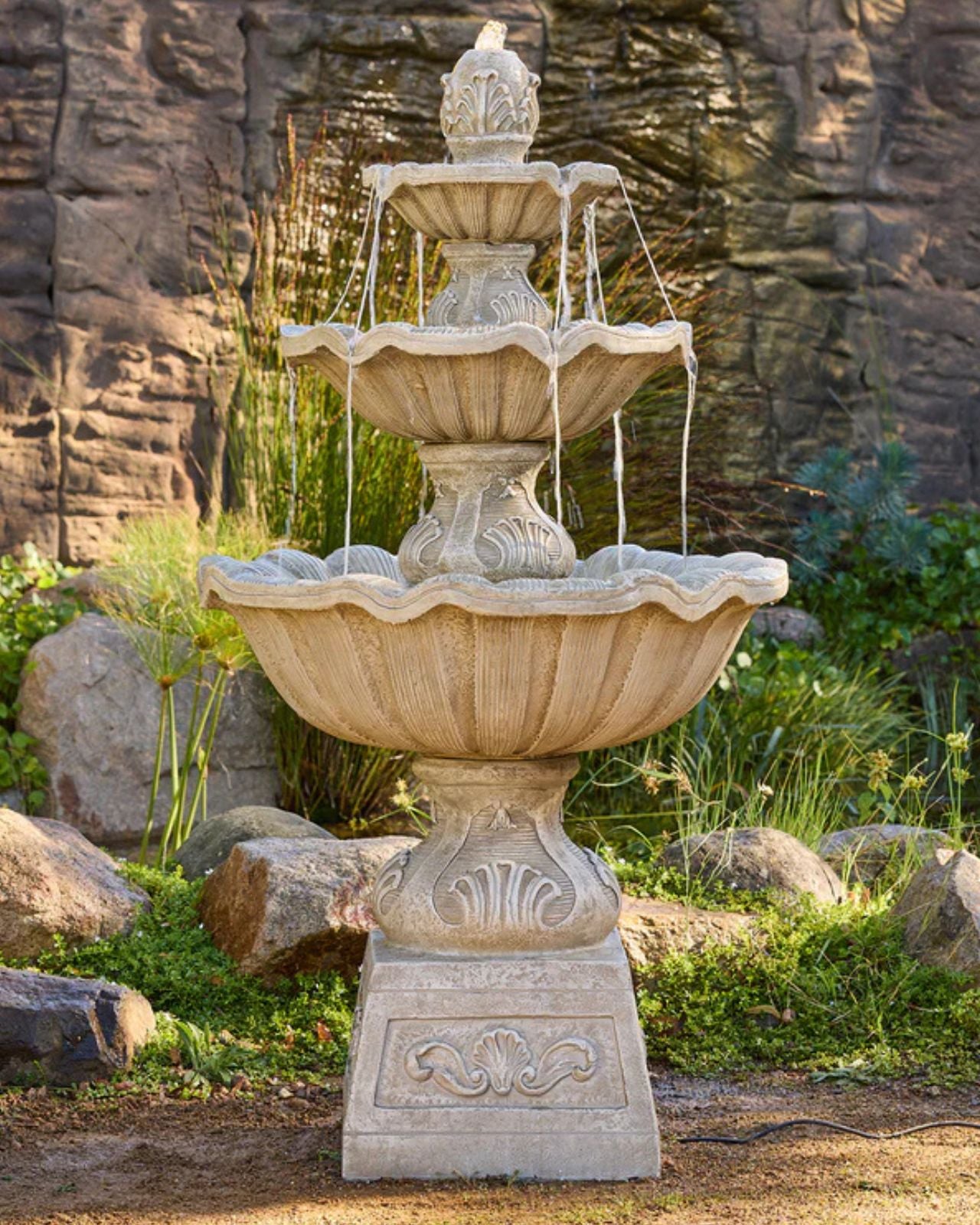 Three-tiered stone fountain in a garden setting with rocks and plants.