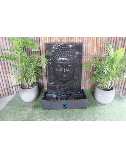 Fountain with Buddha face design in a garden setting with plants and a wooden fence.