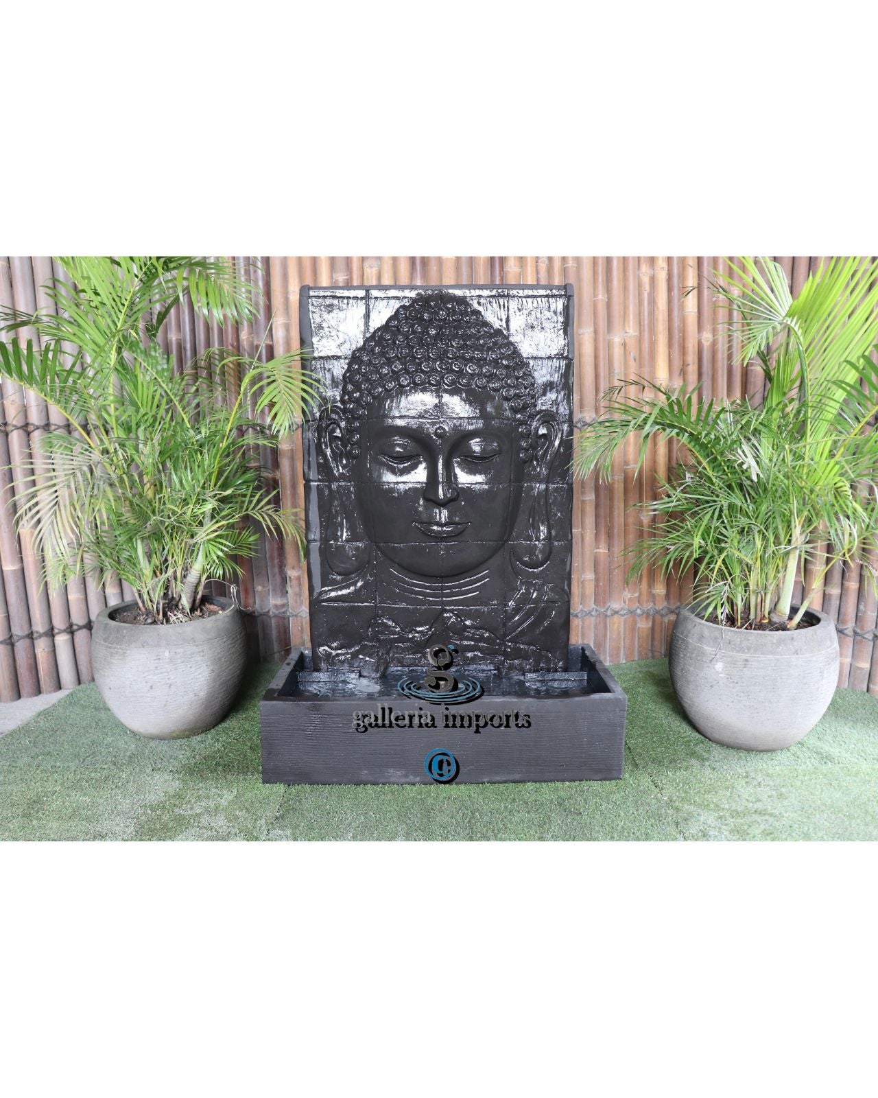 Decorative water fountain with Buddha face design in a garden setting with plants and wooden fence.