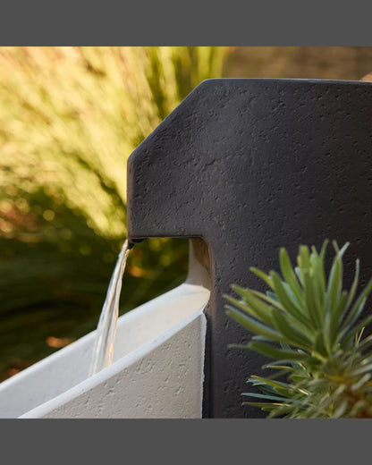 Aegea water feature spout with water flowing into a white basin, surrounded by greenery.