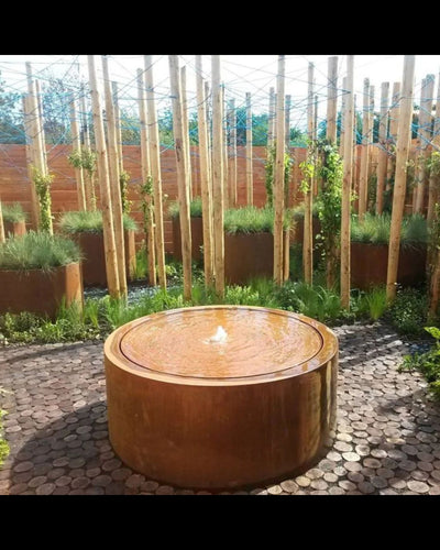 Round metal fire pit water feature in a garden setting with plants and wooden fence.