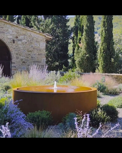 Cylindrical corten steel water feature in a garden with stone building and trees in the background