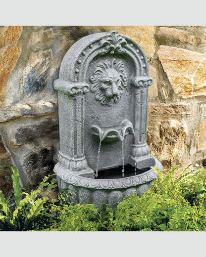 Decorative stone water fountain with lion head design against a stone wall.